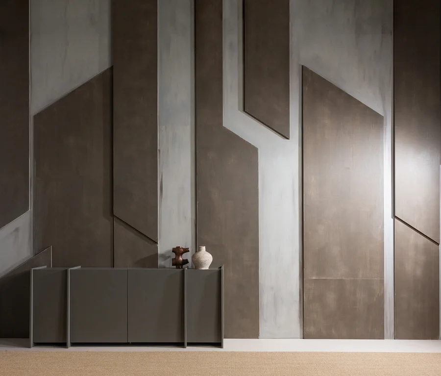 Moderner Raum mit einem dunkelgrauen Sideboard vor einer Wand mit abstrakten geometrischen Paneelen in Dunkel- und Hellgrau. Ein hellbrauner, strukturierter Teppich bedeckt den Boden. Auf dem Sideboard stehen eine dunkle Holzskulptur und eine helle Vase.