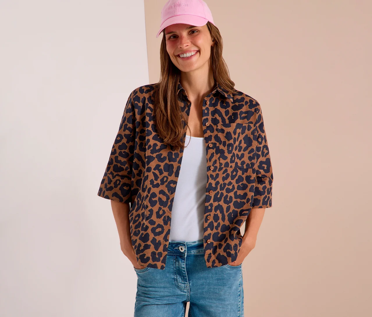 Lachende Frau mit pinker Baseballkappe, offener CECIL Hemdbluse mit Leopardenmuster über weißem Top und hellblauer Jeans, Hände in den Taschen.