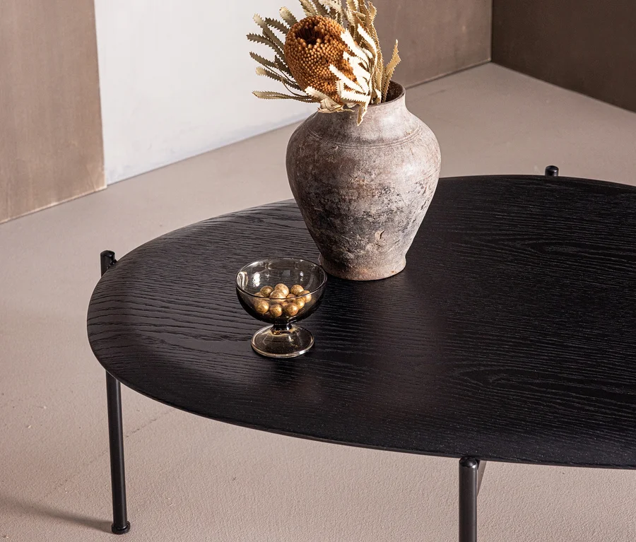 Schwarzer ovaler Couchtisch mit einer rustikalen Vase, die Trockenblumen enthält, und einer kleinen Glasschale mit goldenen Dekokugeln.