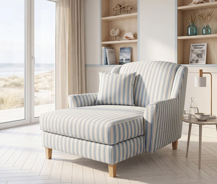Blau-weiß gestreifte Chaiselongue in einem hellen Raum mit Blick auf einen Sandstrand und das Meer. Daneben stehen ein Beistelltisch mit Lampe und Regale mit Büchern und Muscheln.