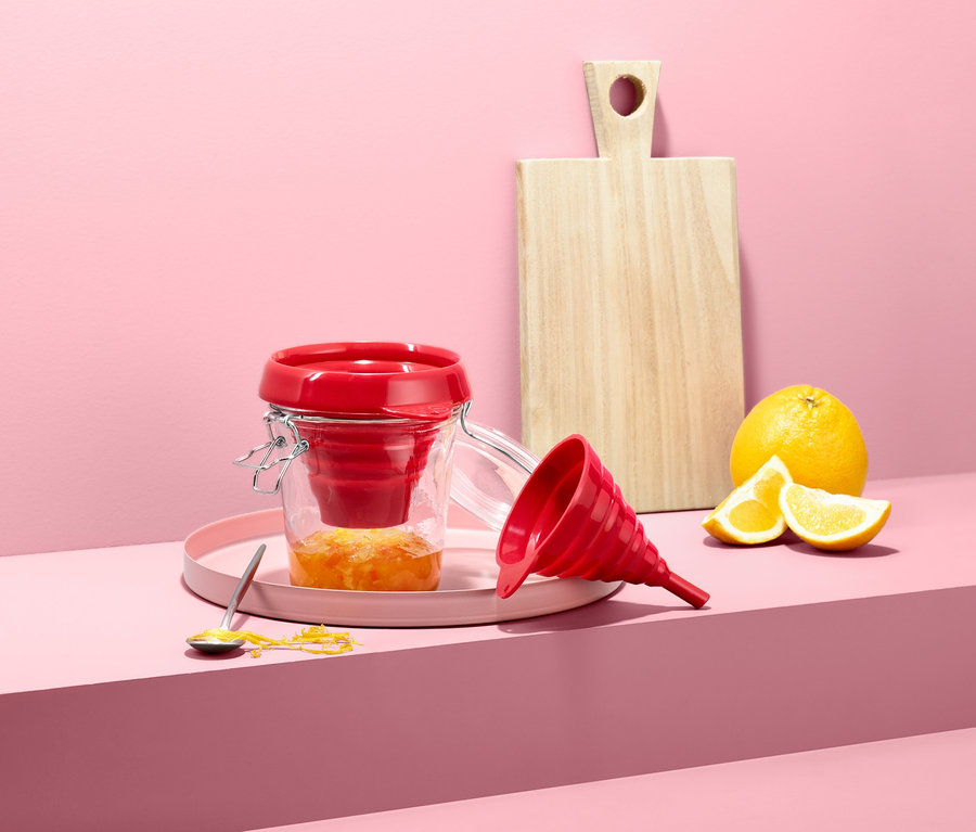 Ein roter Silikon-Trichter befindet sich in einem offenen Glas mit Marmelade, ein zweiter Trichter liegt daneben, auf rosafarbenem Hintergrund mit Brett und Zitrusfrüchten.
