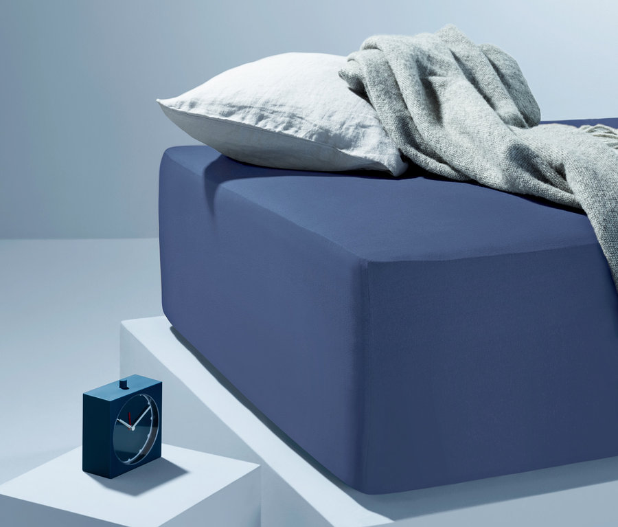 Blaues Bett mit weißem Kissen und grauer Decke auf weißen Würfeln. Daneben steht ein blauer Wecker. Extrahohe Jersey-Spannbettlaken in Normalgröße, Doppelgröße und Übergröße sind verfügbar.