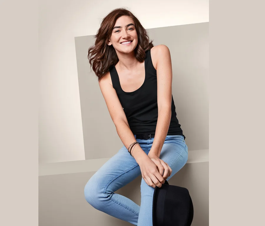 Eine lächelnde Frau sitzt in Jeans und einem schwarzen Top. Sie hält einen schwarzen Hut. Sie trägt 2 Tops.