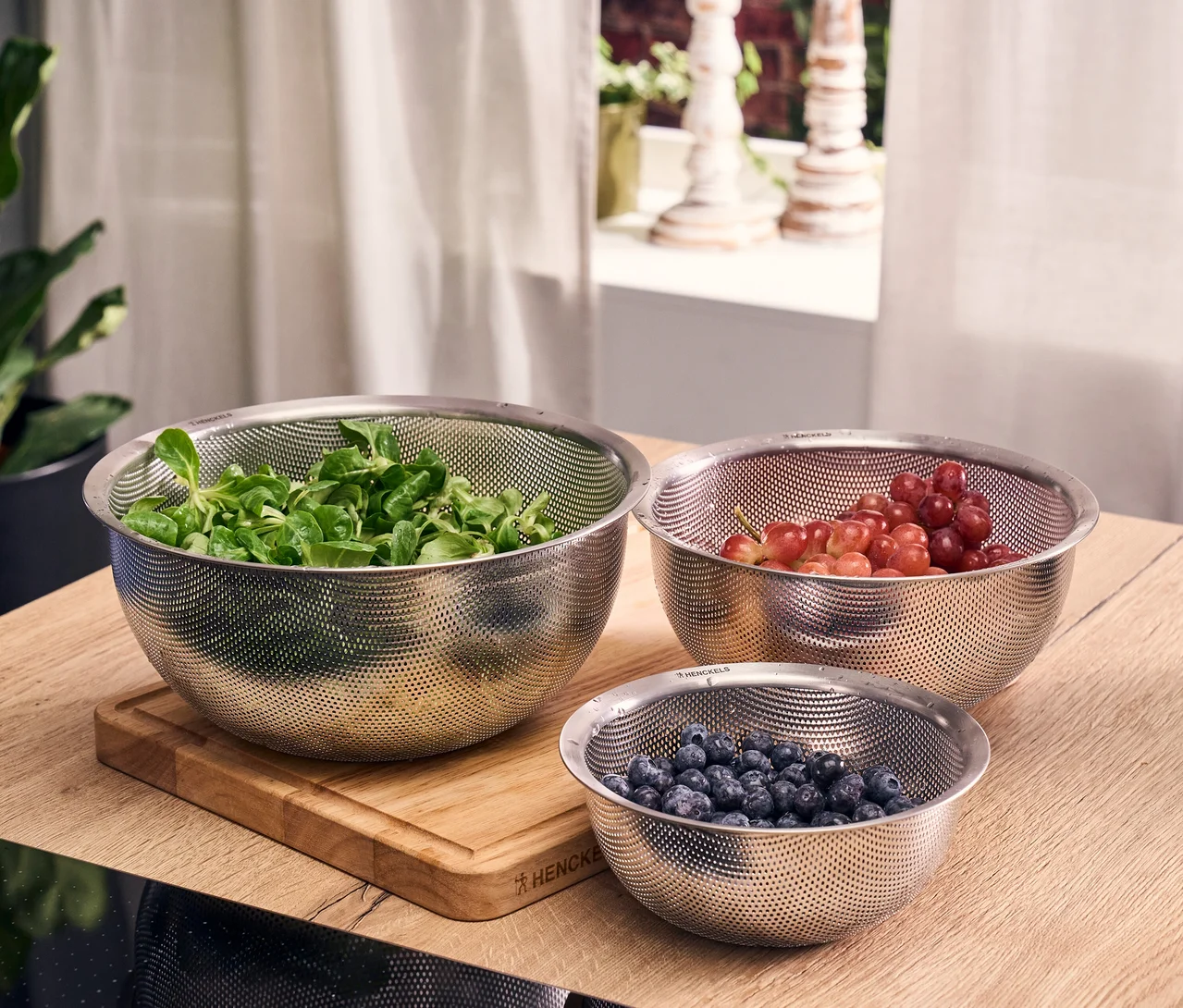 Drei Edelstahl-Siebe aus dem Henckels by ZWILLING 3-teiligen Siebset auf einer Holztheke. Das größte enthält Feldsalat, das mittlere rote Weintrauben und das kleinste Heidelbeeren.