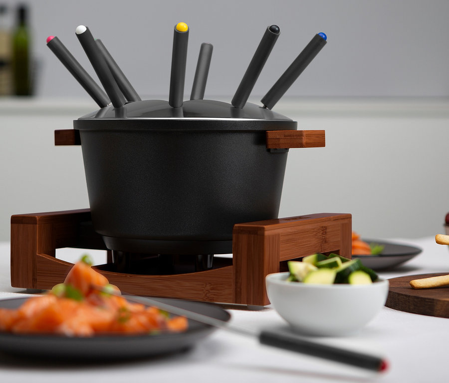 Schwarzes Fondue steht auf einem Holzständer, umgeben von Tellern mit Essen und Fonduegabeln.