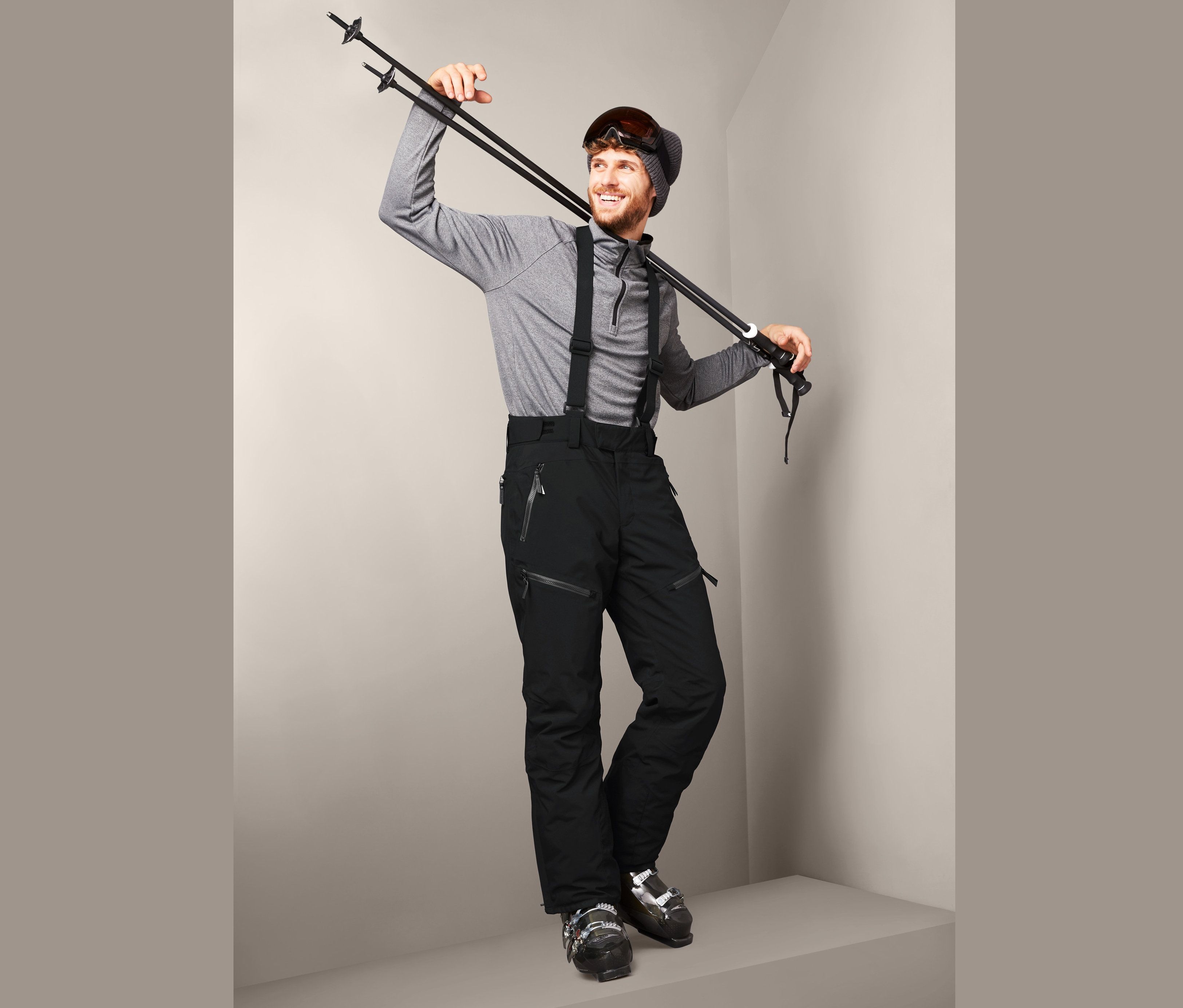 Mann posiert mit Skistöcken über der Schulter, trägt eine schwarze High-End-Skihose und Skischuhe.