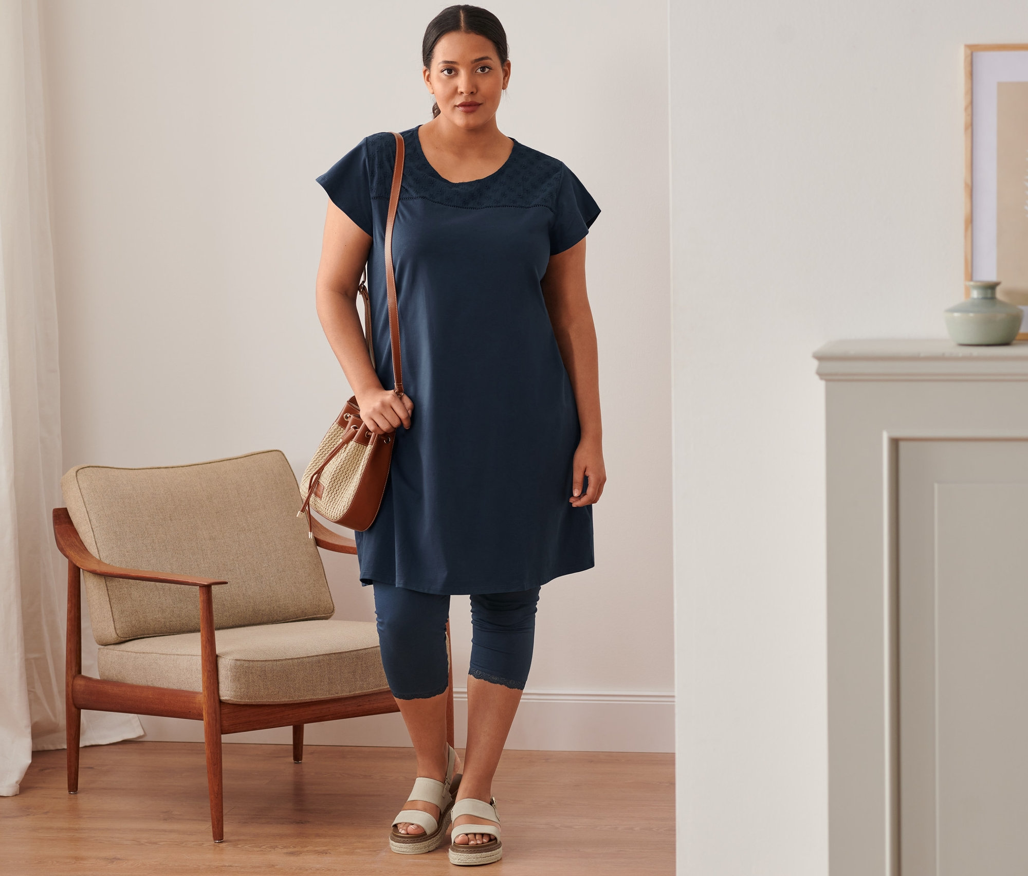 Frau steht und trägt ein marineblaues Jerseykleid mit Lochstickerei, 3/4-Leggings mit Spitzenborte, Plateau-Sandale aus Leder und eine Beuteltasche.