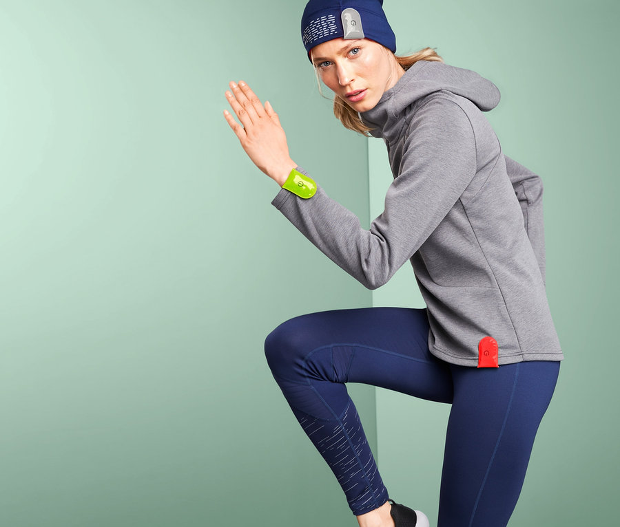 Frau in grauem Kapuzenpullover und blauen Leggings mit 3 LED-Magnetlichtern am Arm und an der Mütze.