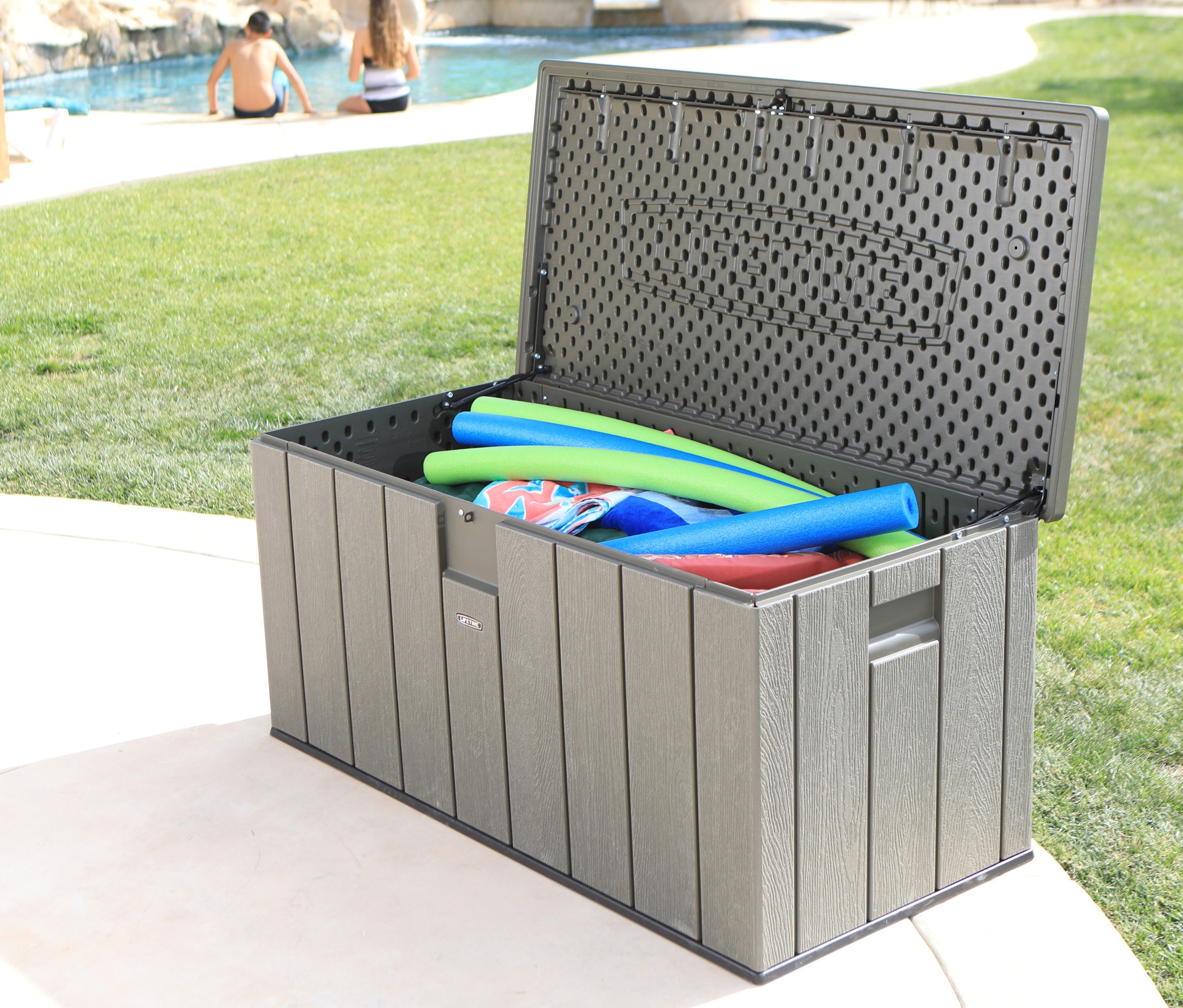 Geöffnete Auflagenbox (ca. 570 l) mit Schwimmnudeln auf einer Terrasse am Pool, im Hintergrund Personen am Pool.