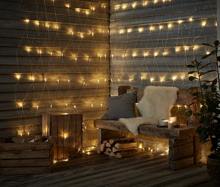 Eine Holzbank mit Kissen und Fell steht vor einer Wand, die mit einem LED-Outdoor-Lichtnetz geschmückt ist.