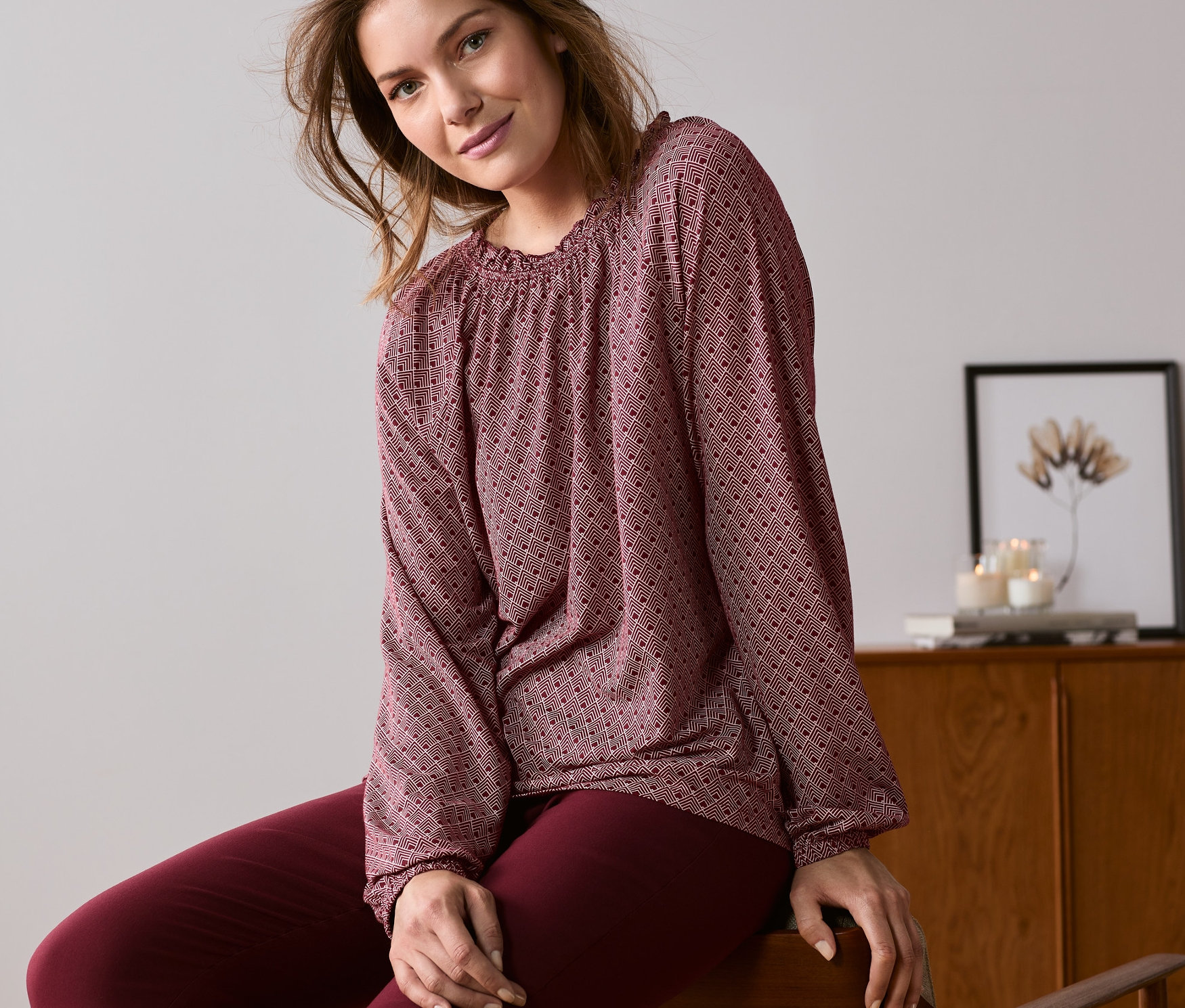 Frau sitzt auf einem Stuhl und trägt ein dunkelrotes, bedrucktes Blusenshirt.
