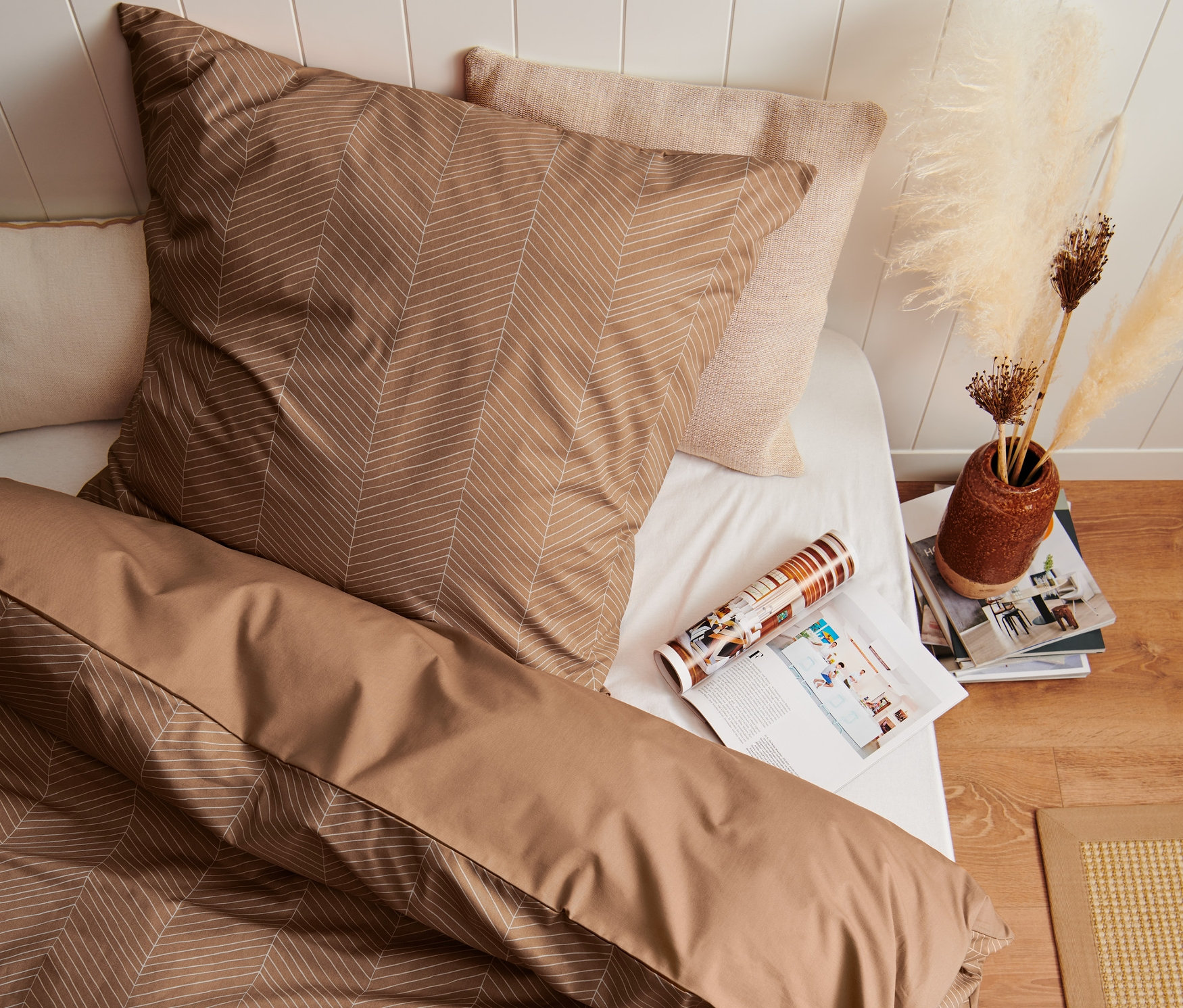 Draufsicht auf ein Bett mit beiger Premium-Baumwoll-Bettwäsche mit geometrischem Muster, daneben eine Zeitschrift und eine Vase mit Pampasgras.