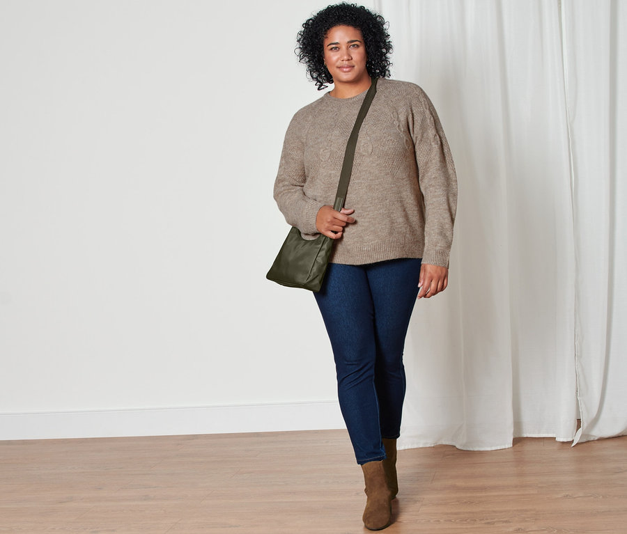 Frau geht, trägt Grobstrick-Pullover mit Zopfmuster, Jeggings, Stiefelette aus Veloursleder und Umhängetasche.