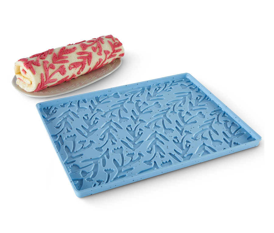 Biskuitrolle liegt auf Teller neben blauer Dauerbackmatte für Biskuitrollen mit Blumenmuster.