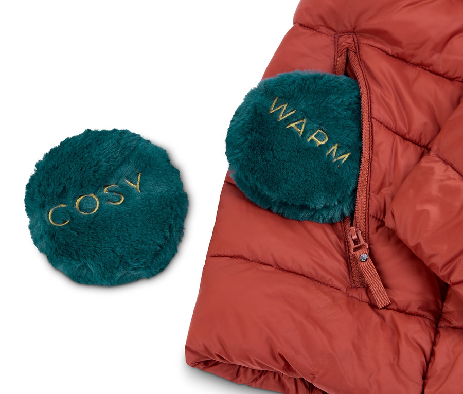 Eine rote Daunenjacke mit 2 Taschen-Heizkissen. Ein Kissen liegt außerhalb der Jacke und trägt die Aufschrift "COSY", das andere befindet sich in der Jackentasche und trägt die Aufschrift "WARM".