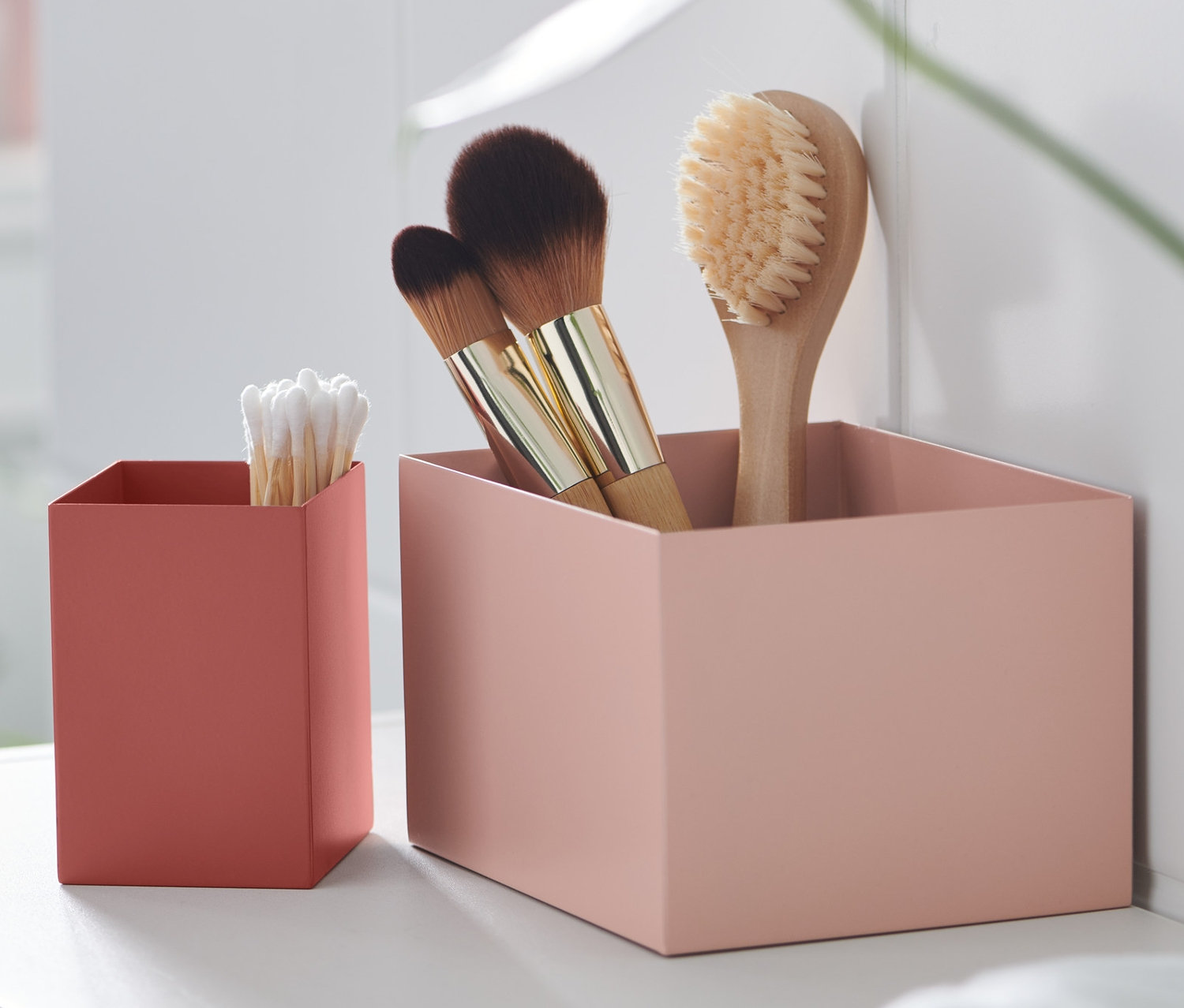 Zwei dekorative Boxen aus Metall stehen auf einem weißen Regal, eine mit Bürsten und die andere mit Wattestäbchen.