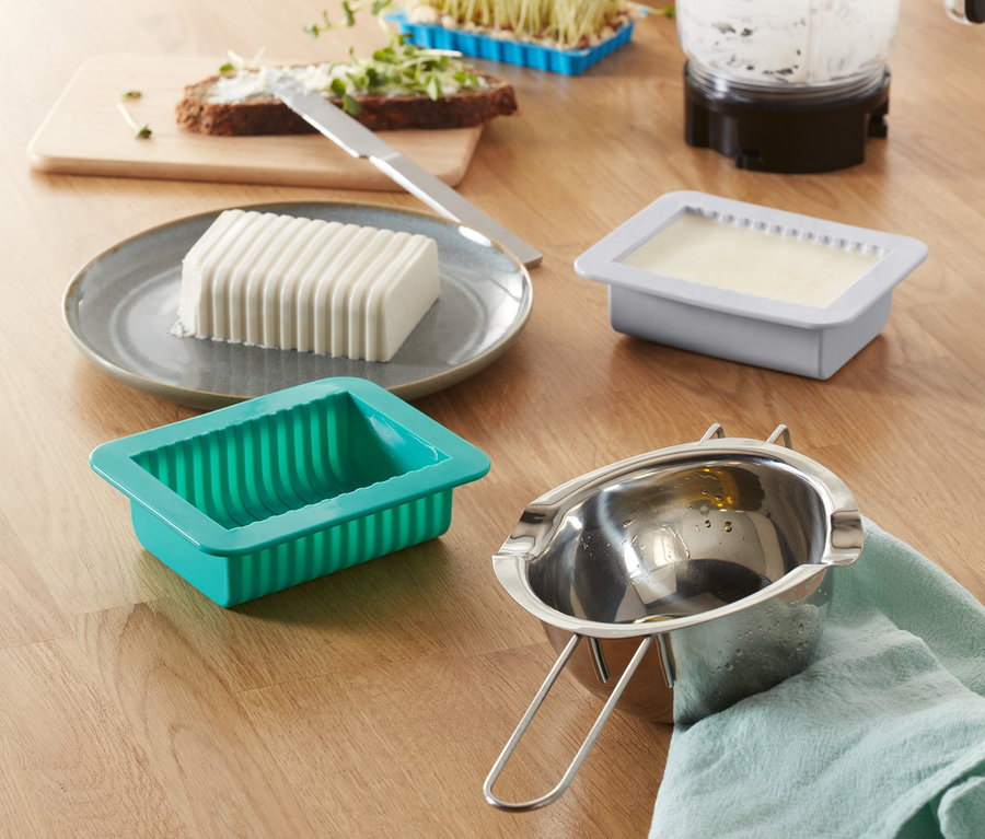 Auf einem Tisch stehen ein DIY-Pflanzenbutter-Set, eine Butterform, ein Mixer, Sprossen und Brot mit Butter.