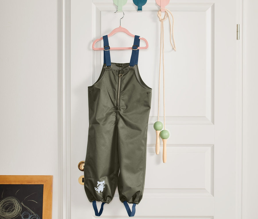 Eine grüne Regenhose hängt an einem rosa Kleiderbügel an einer weißen Tür.