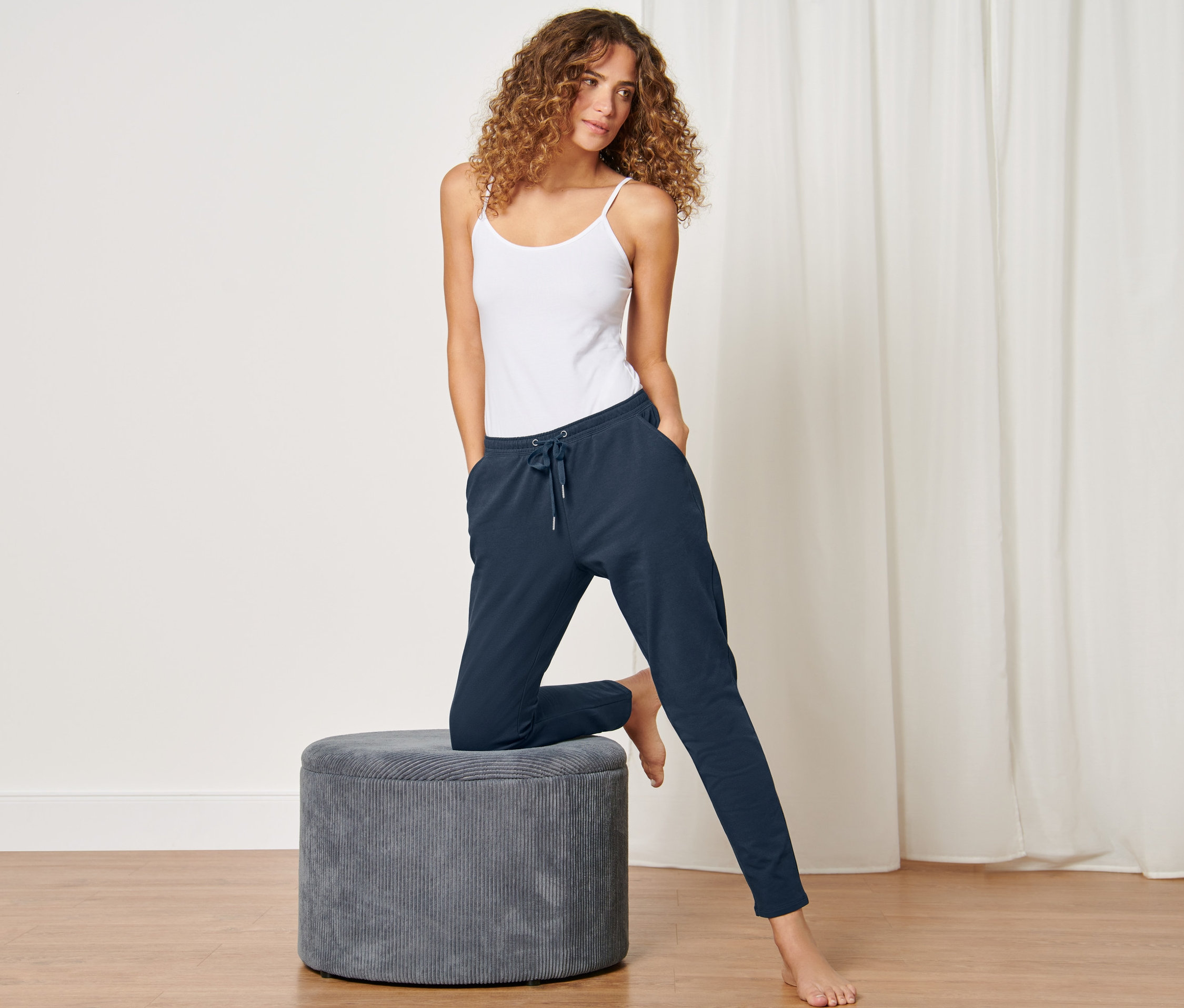 Frau posiert in dunkelblauer Loungewear-Hose und weißem Tanktop, ein Bein auf einem grauen Hocker.
