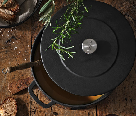 Draufsicht auf einen Holztisch mit einer Suppe in einer Schüssel auf einem Stoffserviette, COMBEKK Gusseisen-Bräter, Holz-Schneidebrett, Drahtschale, Weingläser und Musselinservietten.