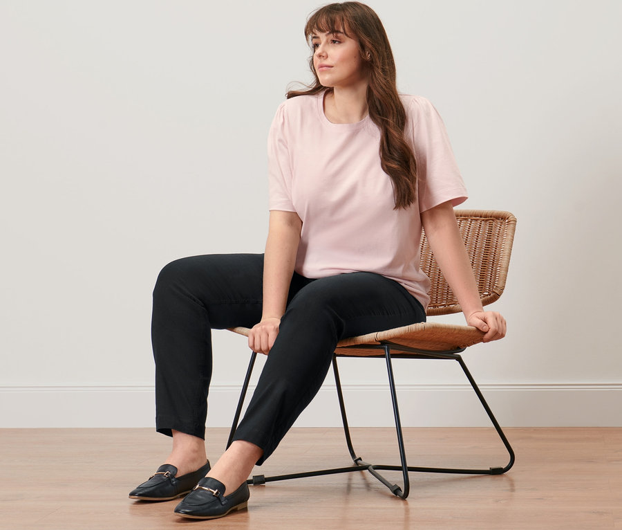 Eine Frau sitzt auf einem Stuhl und trägt ein roséfarbenes Shirt mit Raffung und eine schwarze, konfektionierte Bengalinhose.