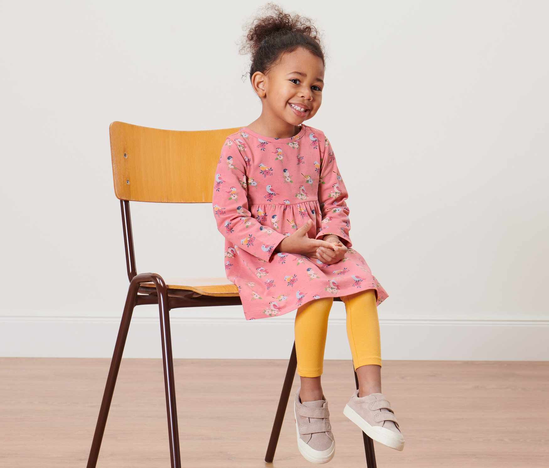 Lächelndes kleines Mädchen sitzt auf einem Stuhl und trägt ein Kleinkind-Jerseykleid.