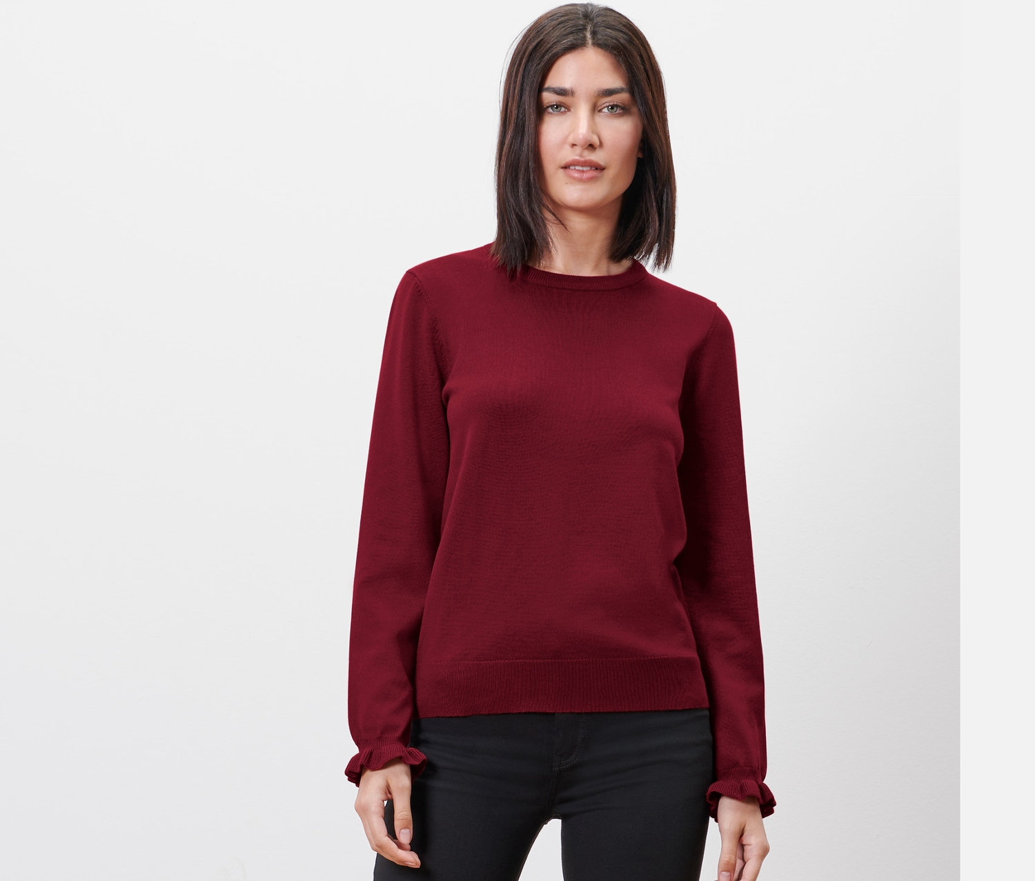 Frau posiert in einem bordeauxroten Feinstrickpullover mit Rüschendetail und einer schwarzen Hose.