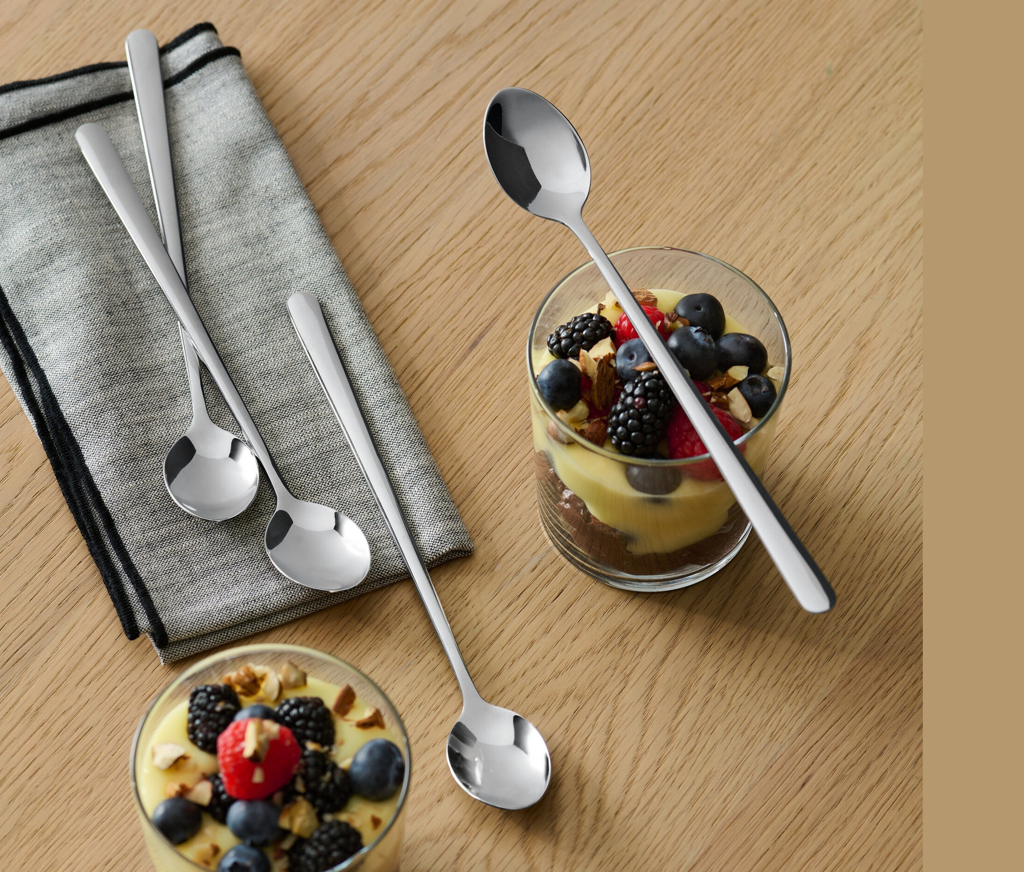 Auf einem Holztisch liegen ein graues Tuch und zwei Gläser mit Desserts und Früchten. Neben jedem Glas liegt einer von 4 langen Löffeln.