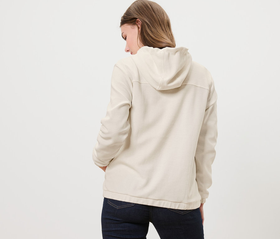 Rückansicht einer Frau mit den Händen in den Hosentaschen, die eine cremeweiße Sweatjacke trägt.