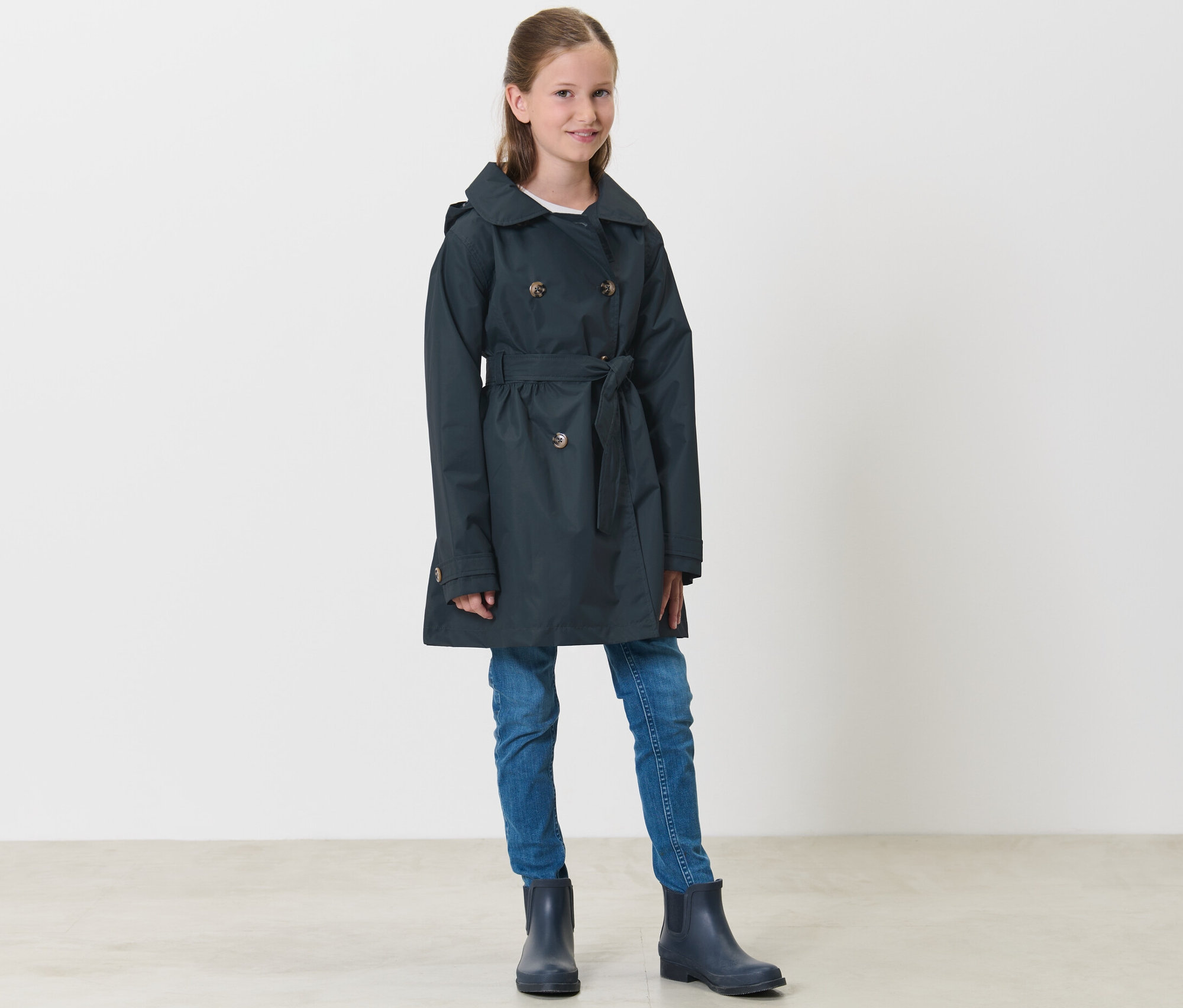 Mädchen steht und trägt einen marineblauen Kinder-Allwetter-Trenchcoat, Jeans und Gummistiefel.