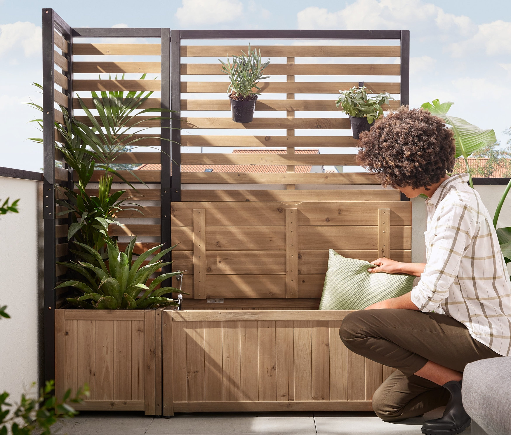 Eine Frau legt ein Kissen auf ein Sichtschutz-Seiten-Modul »Elin« mit Stauraum-Bank. Im Hintergrund befindet sich ein Sichtschutz-Eck-Modul »Elin« mit Pflanzkasten.