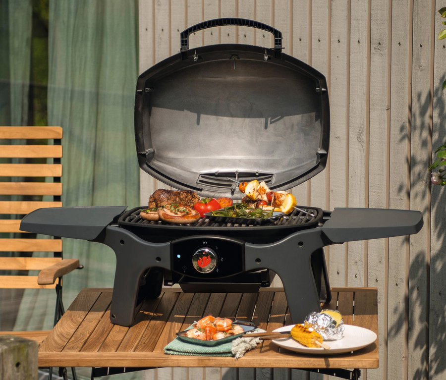 Auf einem Tisch steht ein Grill mit offenem Deckel, auf dem Essen gegrillt wird. Neben dem Grill stehen Teller mit Essen, darunter ein Landmann Elektrogrill »12975 Pantera 2.000 W«.