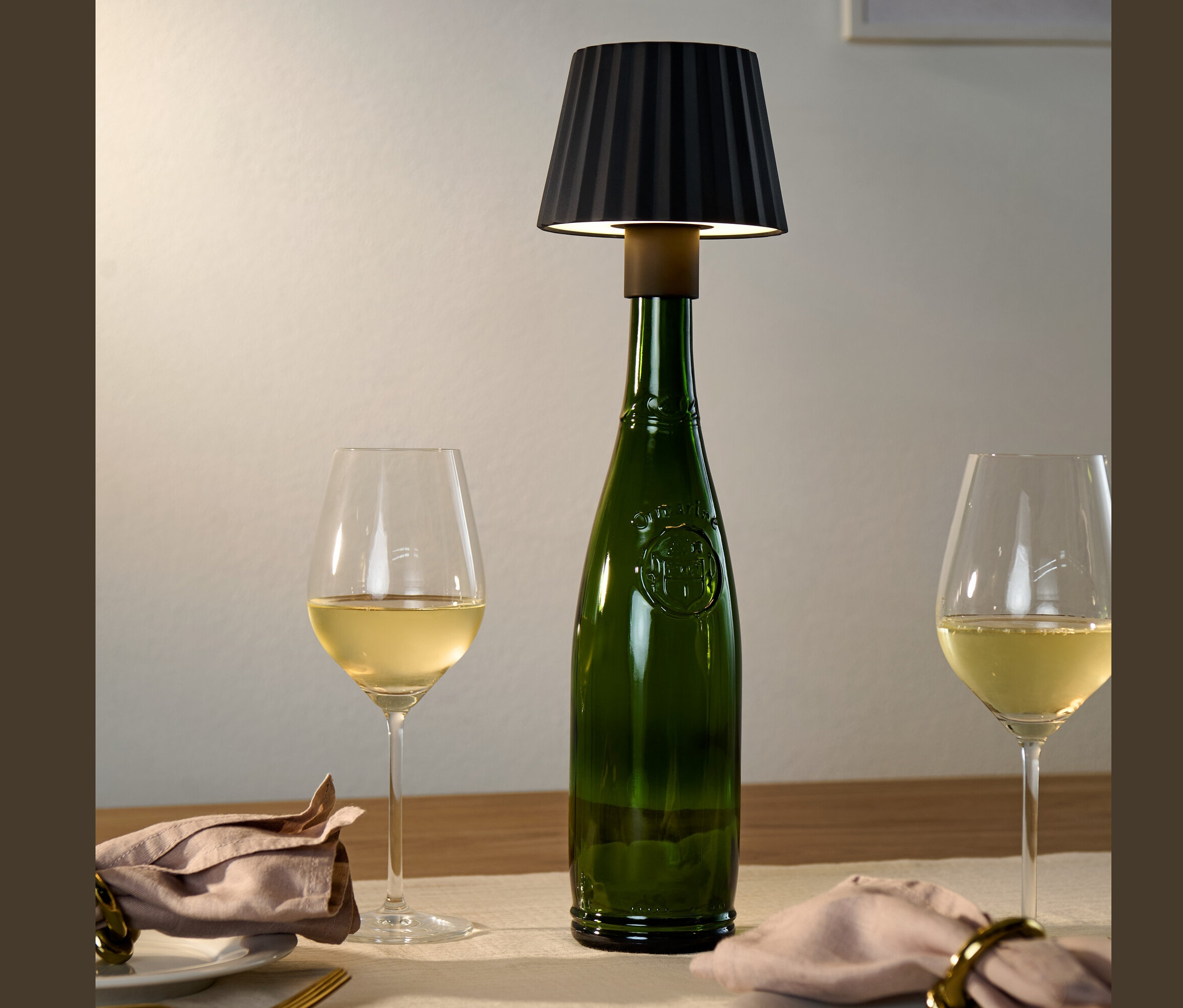 Auf einem Tisch stehen eine grüne Flasche mit einer schwarzen LED-Flaschenleuchte, sowie zwei Gläser Weißwein.