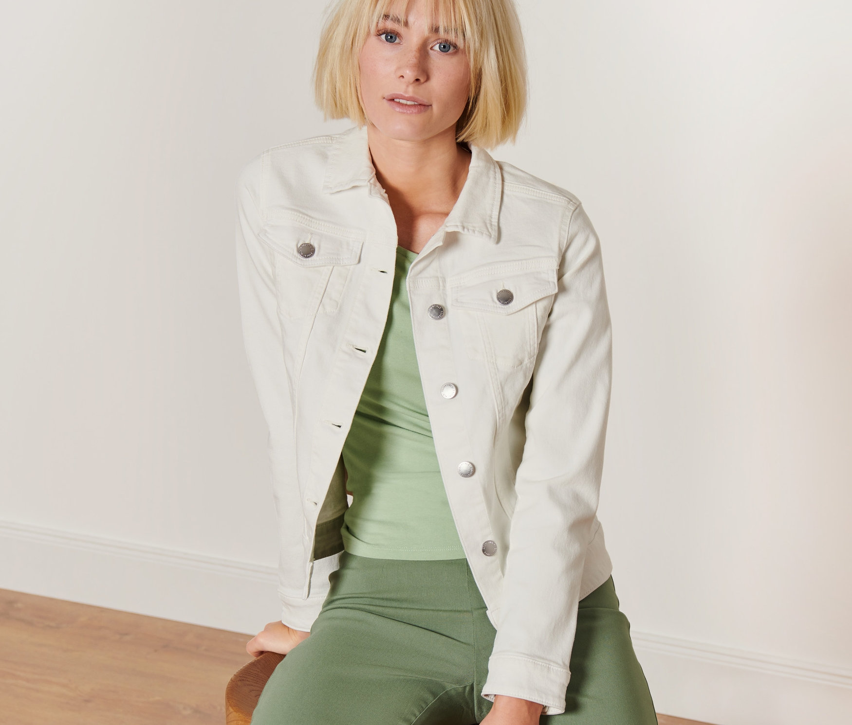 Blonde Frau sitzt auf einem Hocker und trägt eine cremefarbene Jeansjacke.