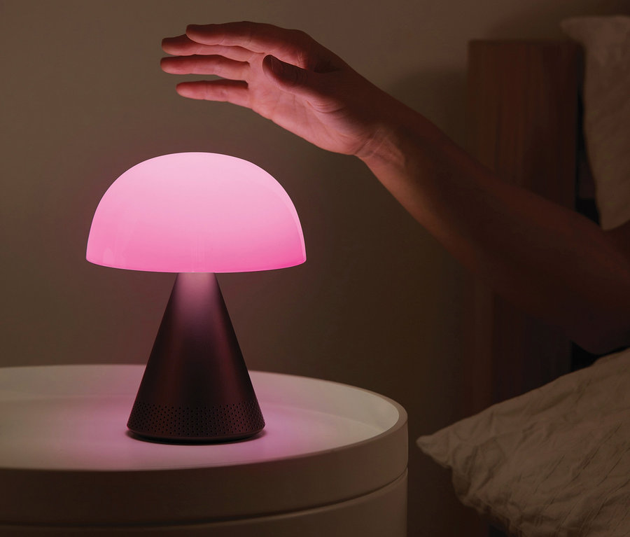 Eine Hand greift nach der anthrazitfarbenen LEXON Akku-Tischleuchte »Mina L Audio«, die auf einem weißen Nachttisch neben einem Bett steht.