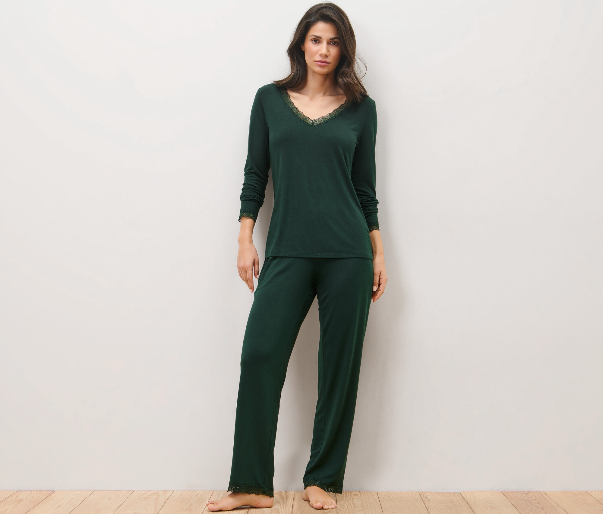 Eine Frau steht auf einem Holzboden und trägt einen dunkelgrünen Pyjama mit Spitzenbesatz.