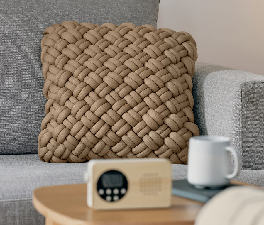Auf einem grauen Sofa liegt ein beiger Strick-Dekokissen-Bezug. Auf einem Holztisch stehen ein FM-Kompaktradio und eine weiße Tasse.
