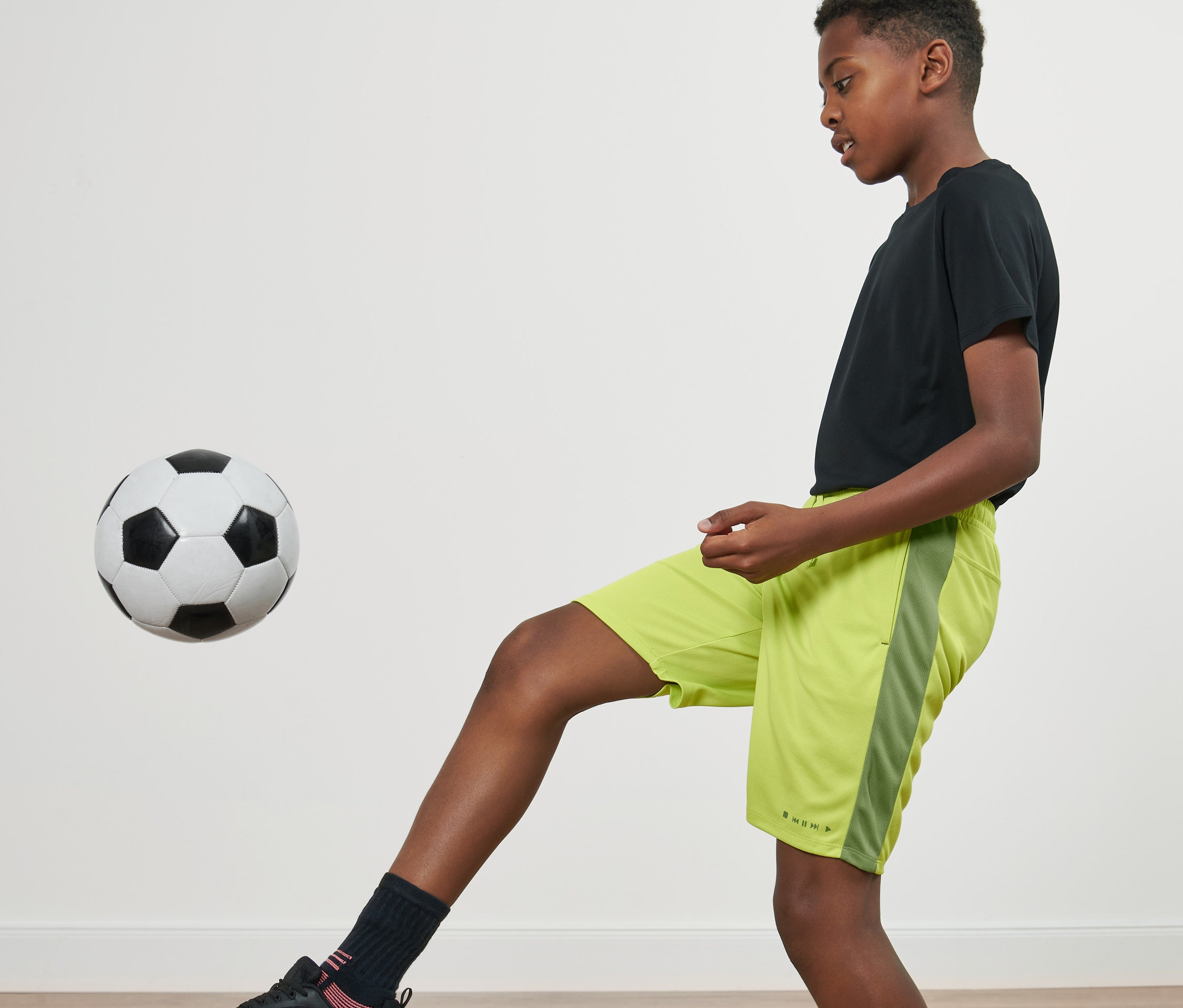 Ein Junge jongliert einen Fußball. Er trägt ein schwarzes T-Shirt und 2 Kinder-Sportshorts.