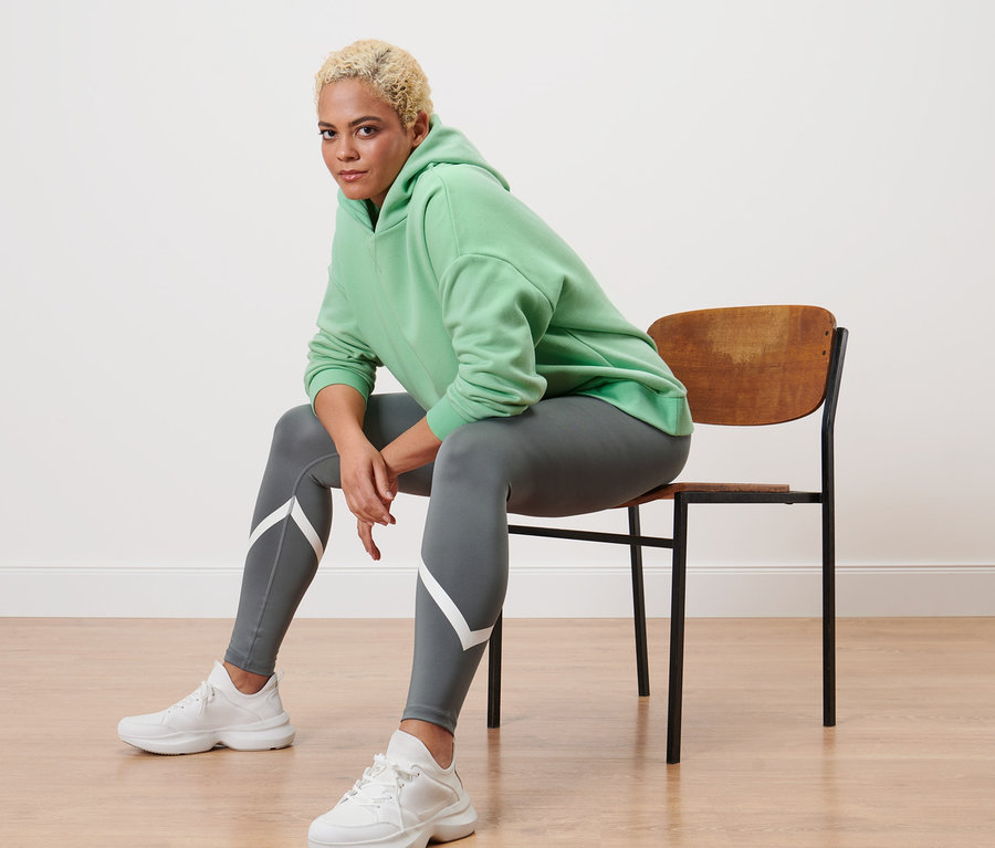 Eine Frau sitzt auf einem Stuhl und trägt einen hellgrünen Hoodie und eine graue Sporttight.