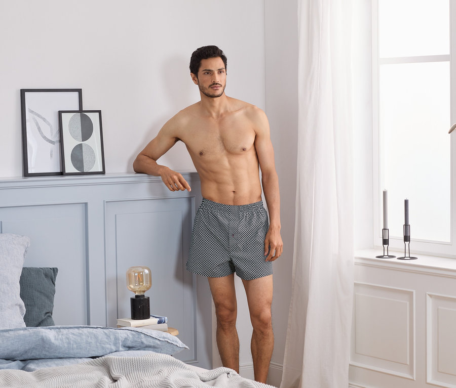Mann lehnt am Bettrahmen und trägt 2 Web-Boxershorts.