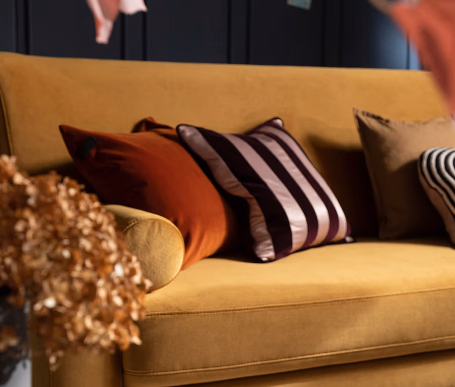 Senfgelbes Sofa mit mehreren Zierkissen, darunter ein rostfarbenes, ein gestreiftes und ein braunes Kissen. Eines der Kissen ist ein SCHÖNER WOHNEN-Kollektion Dekokissenbezug »AVENUE«. Im Vordergrund sind getrocknete Blumen und ein dunkler Beistelltisch zu sehen, im Hintergrund eine dunkle Wandvertäfelung.