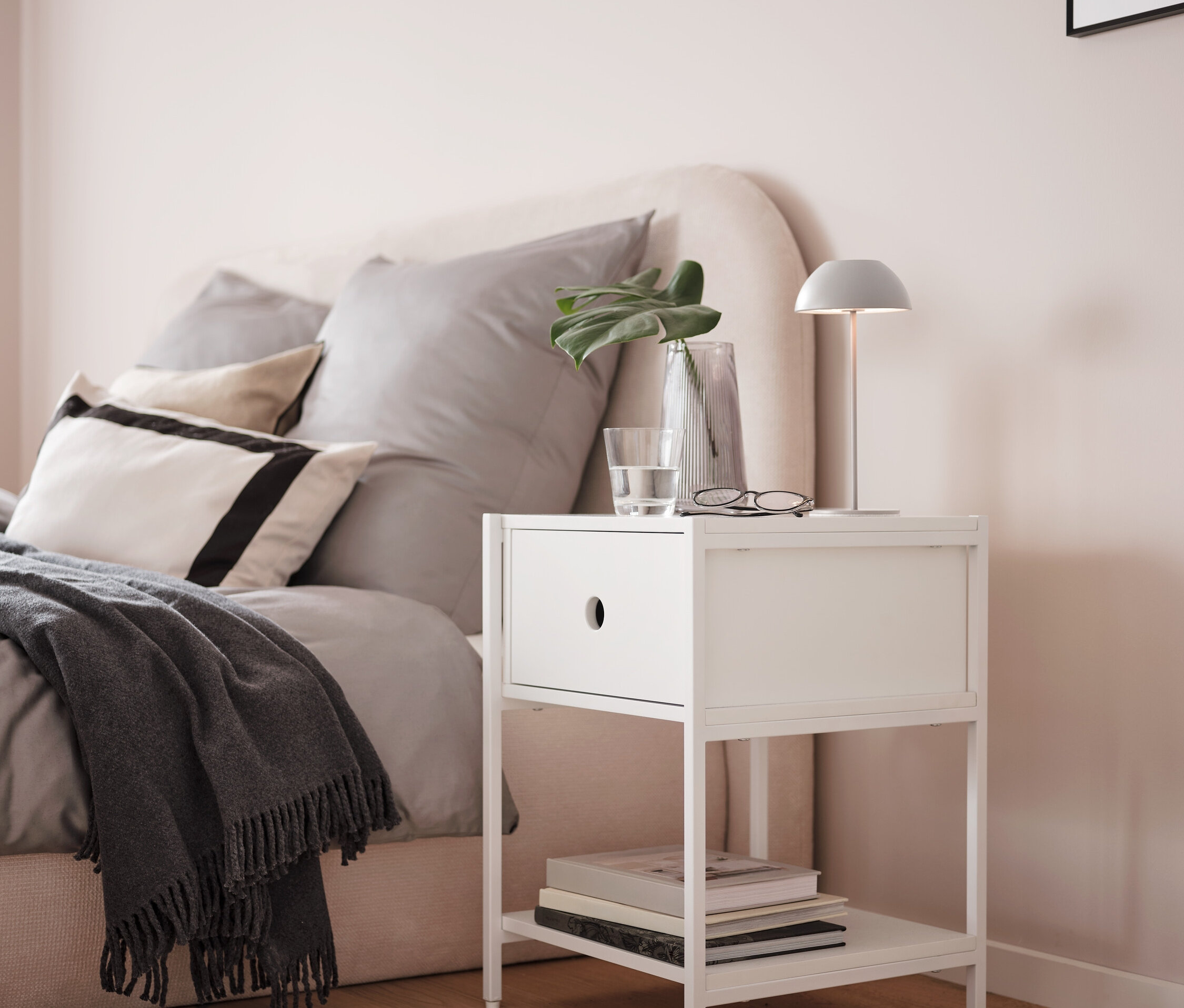 Eine graue LED-Akku-Tischleuchte steht auf einem weißen Nachttisch neben einem Bett.
