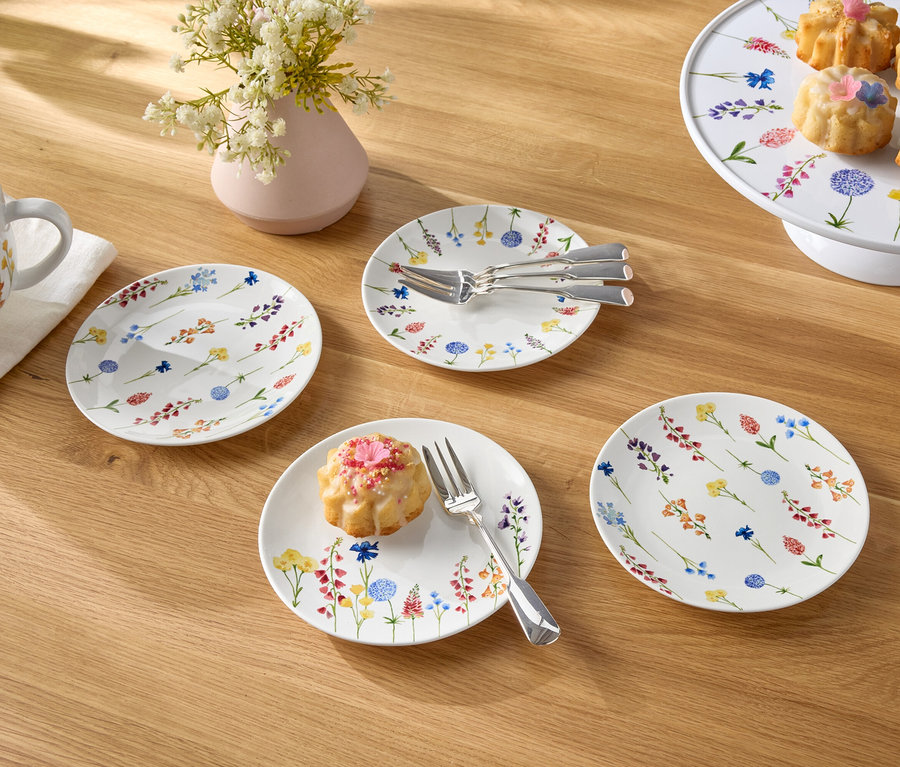 Auf einem Holztisch stehen eine Tasse Kaffee, eine Blumenvase, Teller und kleine Kuchen.