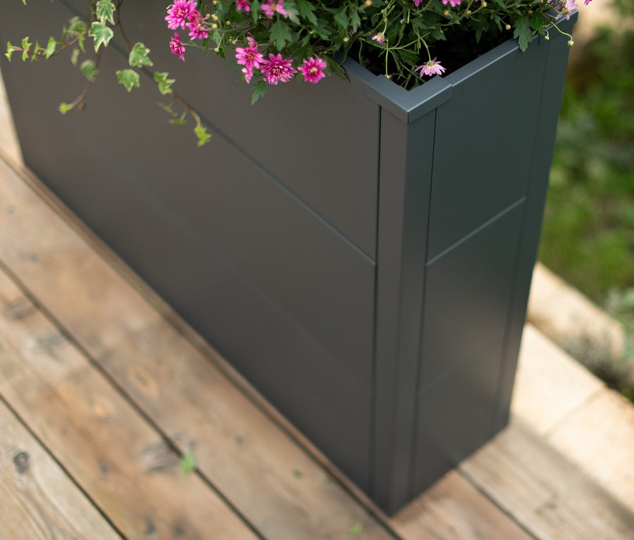 Nahaufnahme eines anthrazitfarbenen Hochbeets mit blühenden rosa Blumen und Efeu auf einer Holzterrasse. Im Bild ist ein ca. 77 cm Pflanzkasten mit Einlegeboden.