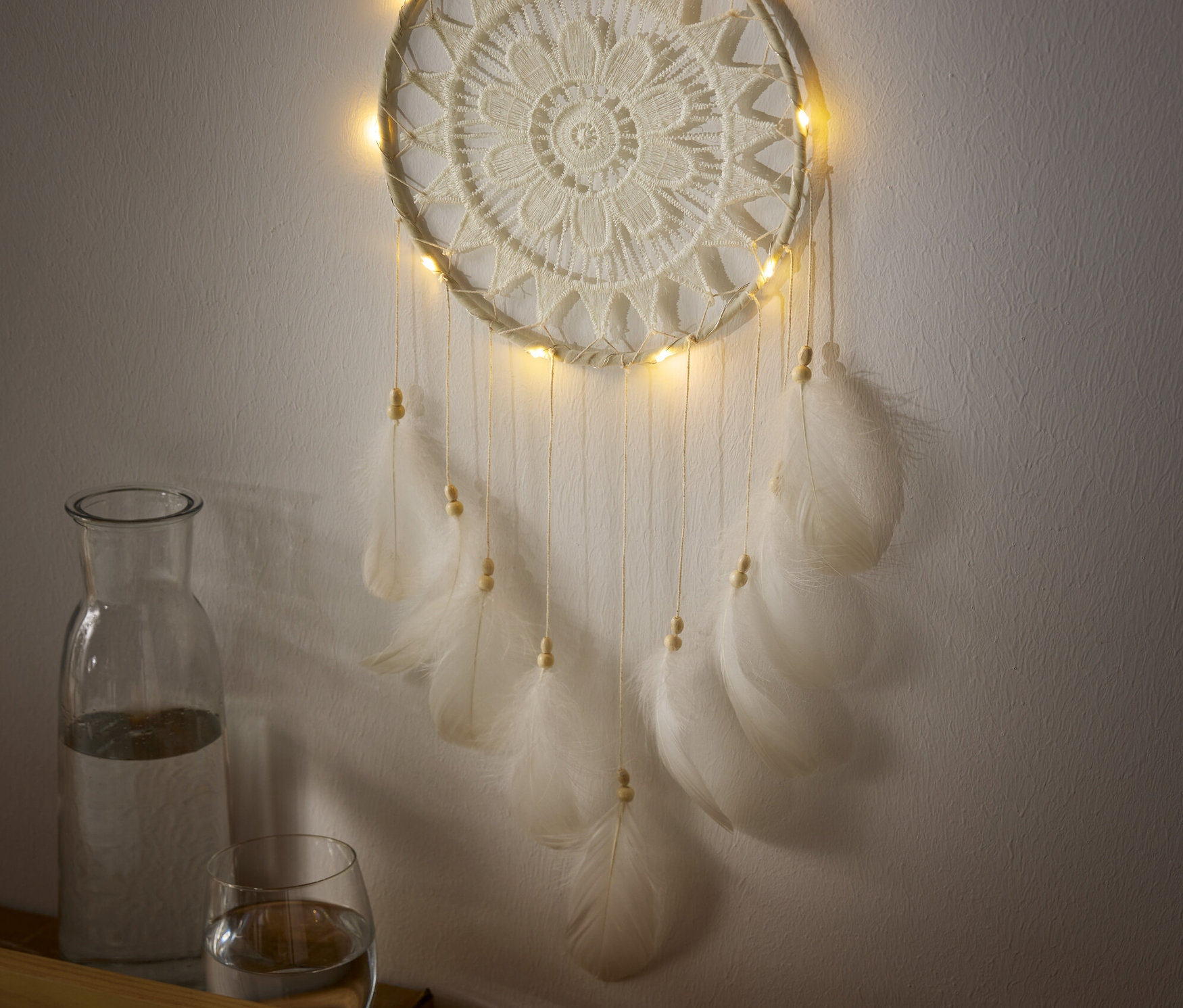 Ein Traumfänger mit LED-Beleuchtung hängt an der Wand, darunter eine Karaffe und ein Glas Wasser.
