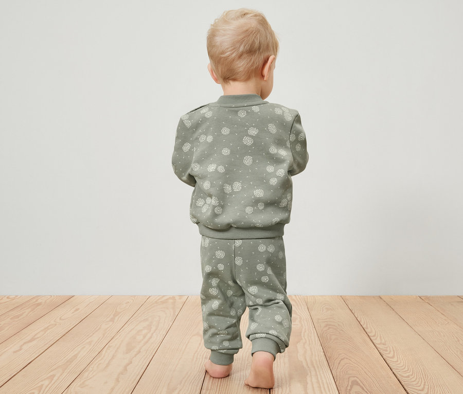 Kleinkind von hinten geht barfuß auf Holzboden und trägt eine grüne Sweat-Kombi.