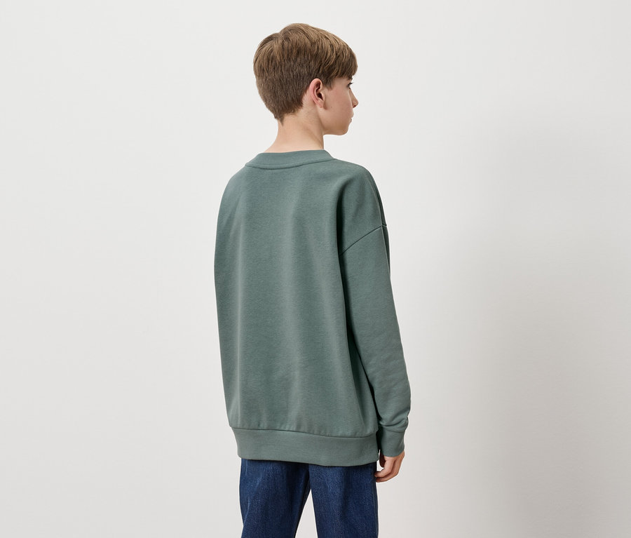 Von hinten ist ein Junge mit kurzen braunen Haaren in einem grünen Sweatshirt und blauen Jeans zu sehen.