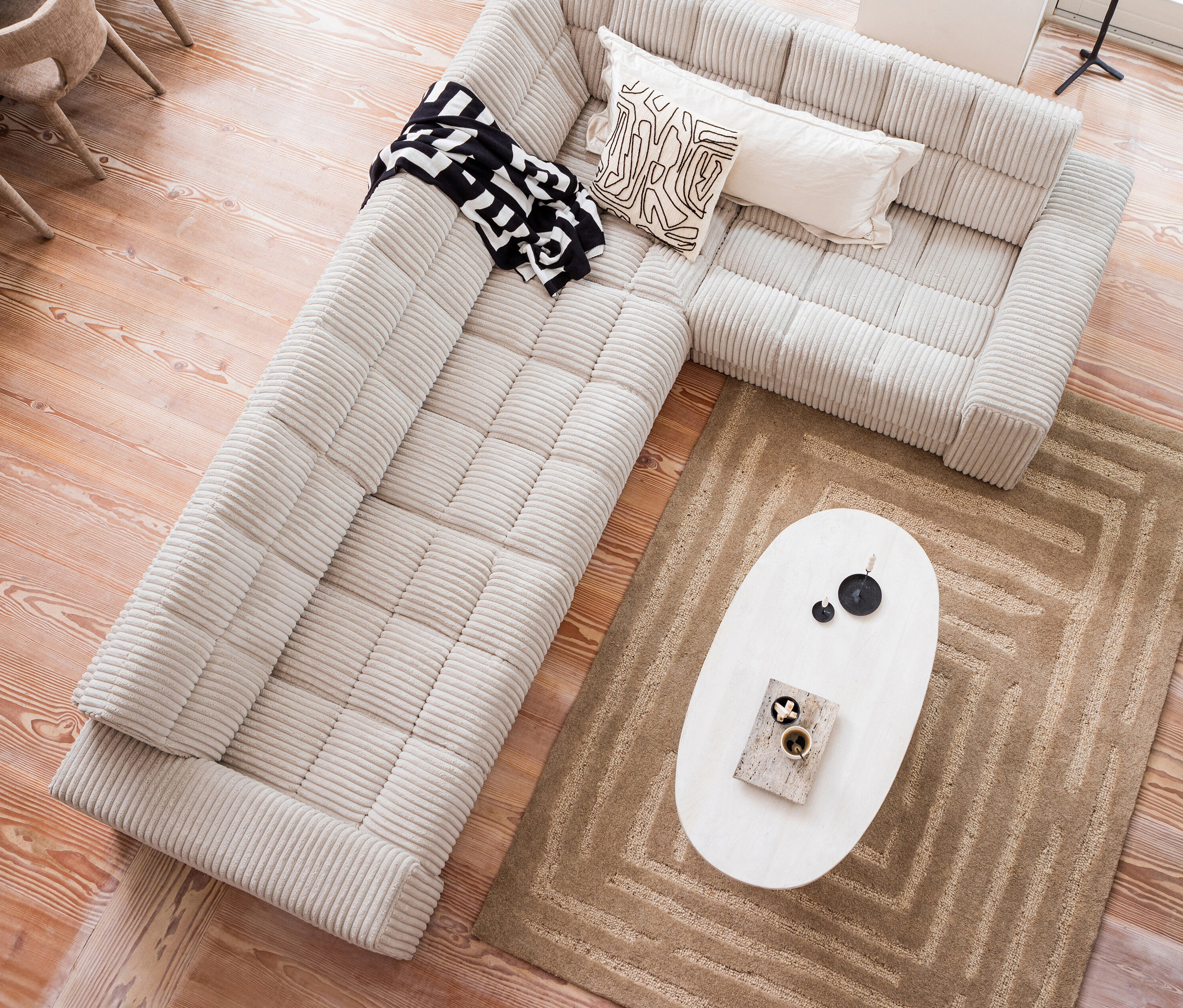 Draufsicht auf ein beiges Ecksofa mit einer schwarz-weissen Decke, zwei dekorativen Kissen und einem weissen Couchtisch mit Dekoration auf einem braunen Woood Teppich Alicia.