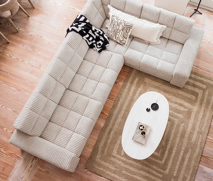 Draufsicht auf ein beiges Ecksofa mit einer schwarz-weissen Decke, zwei dekorativen Kissen und einem weissen Couchtisch mit Dekoration auf einem braunen Woood Teppich Alicia.