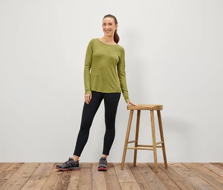 Eine Frau in einem olivgrünen Langarmshirt und schwarzen Leggings steht neben einem Holzhocker.
